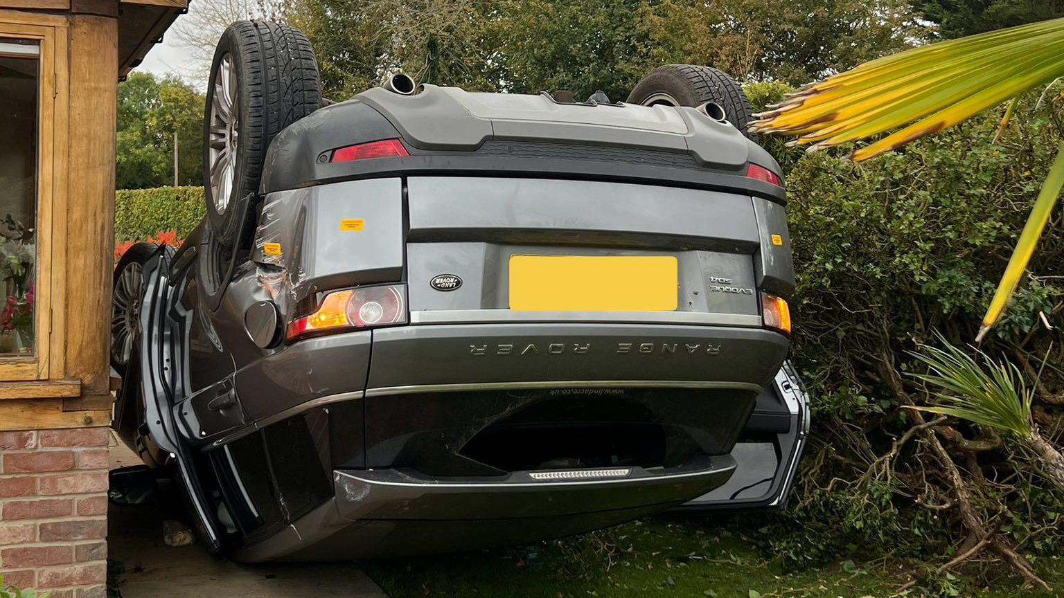 A silver Range Rover on its roof between a brick building on the left, which has brown-stained wooden-framed windows, and a green hedge on the right.
