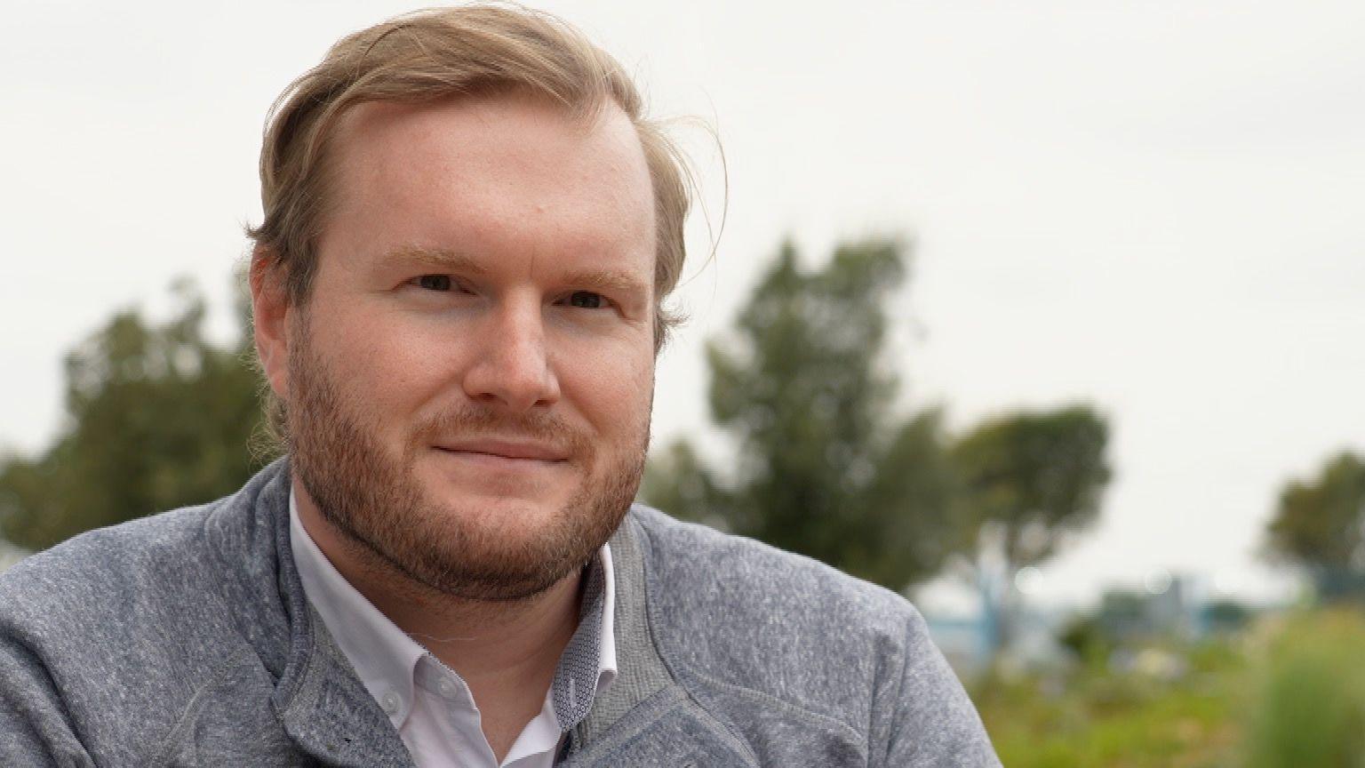 Alexander Sparrow sits in front of trees on a cloudy day. He is wearing a grey jumper with a white collared shirt underneath. He has short hair with a dark, neatly-trimmed beard.