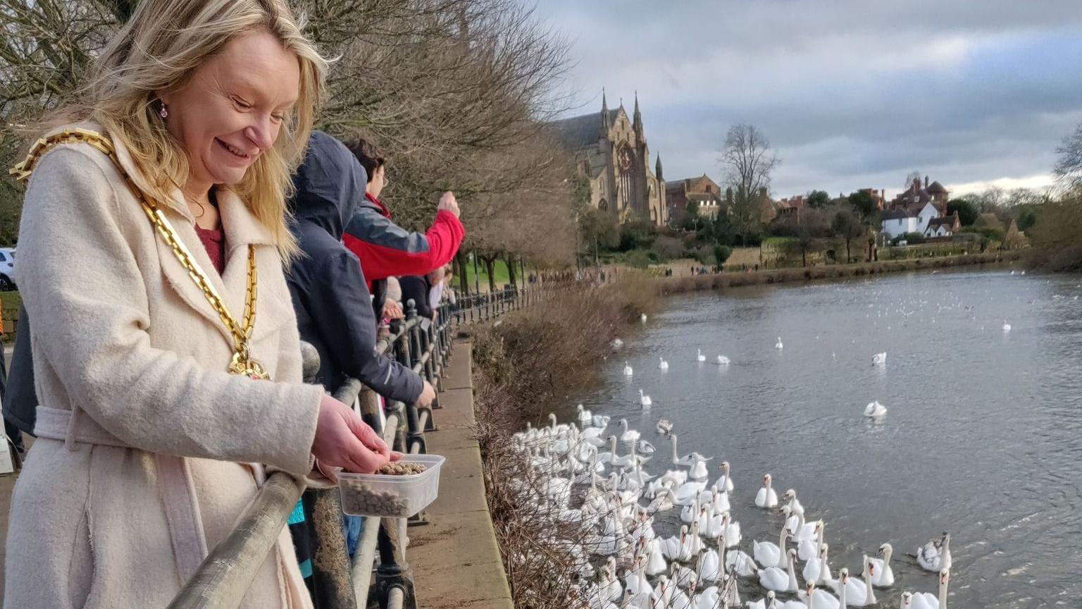 A woman with blonde hair, wearing a cream coat and mayoral chain, stands at a metal railing along the river as she grabs a handful of feed pellets from a plastic tub. Several other people are visible behind her, with dozens of swans gathering nearby in the river.