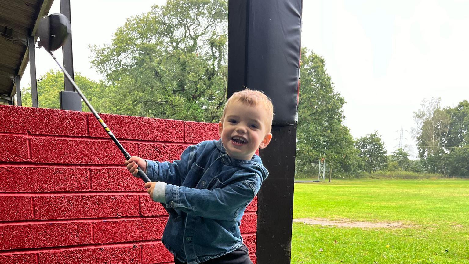 Wilson is mid swing with his new golf club. He is grinning at the camera and holds the driver in the air as he swings it to the right. He has short light brown hair and wears a denim hooded shirt jacket with a grey long sleeve top underneath and black trousers. He is standing in a driving range bay, which has a red brick wall behind him and the green can be seen in the distance, lined by trees.