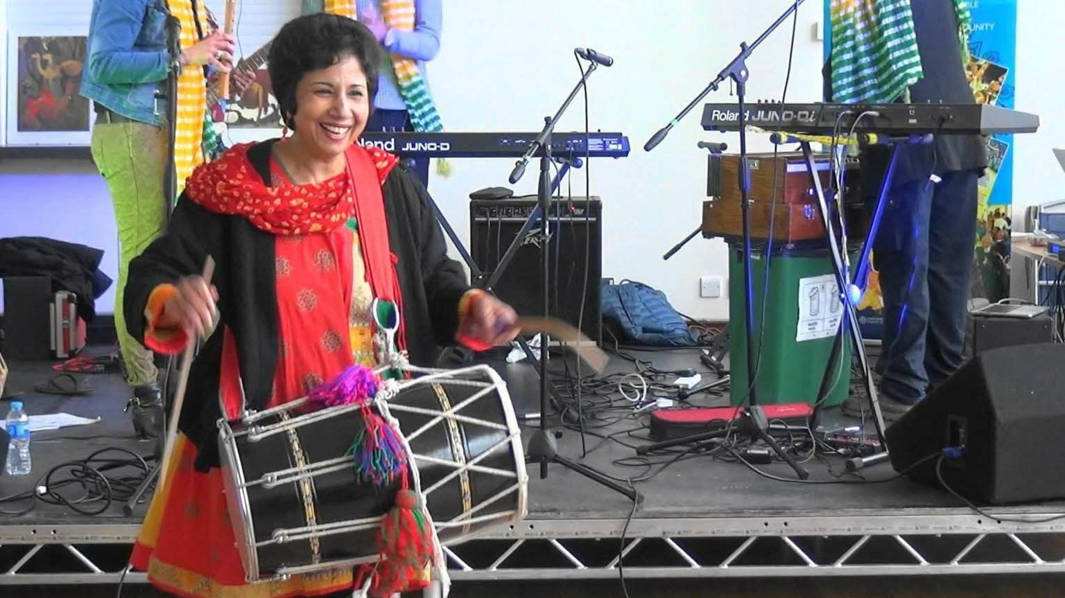 Nuzhat Abbas during a performance. She is smiling while playing drums. She is wearing a colourful outfit. Other musicians can be seen behind her playing guitar and pianos.