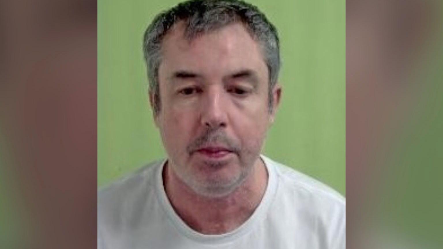 A middle-aged man wearing a white top in a police mugshot 