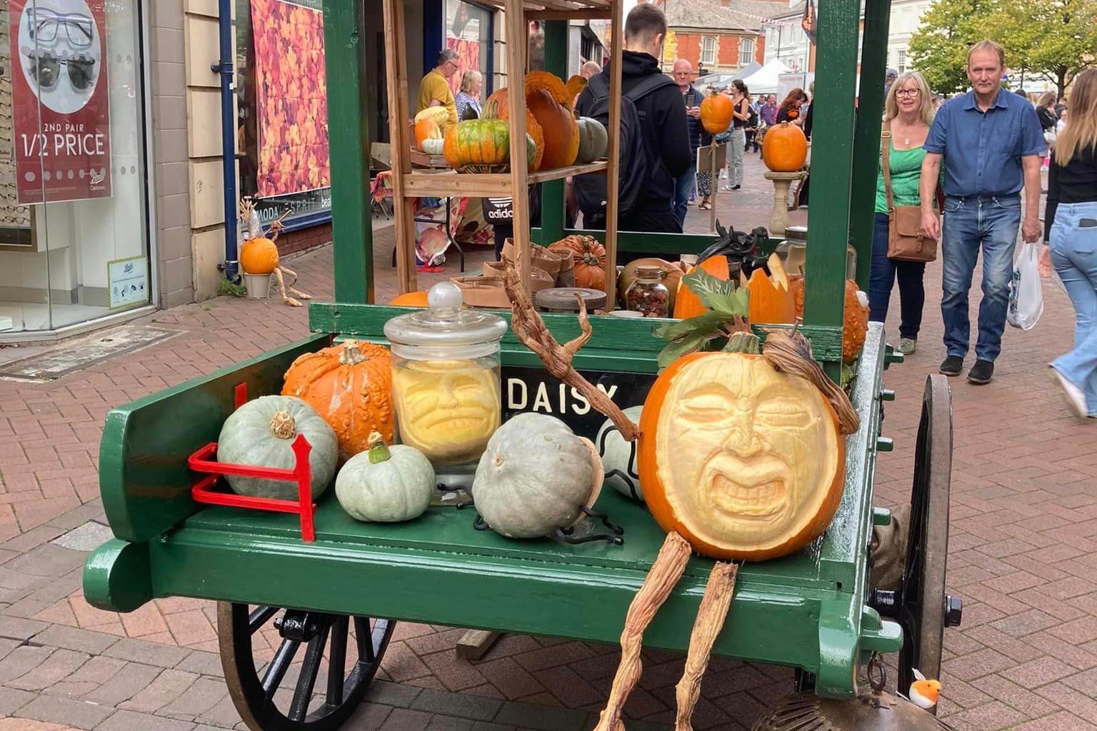 A cart carries carved pumpkins of all kinds on it, as passers-by take a look.