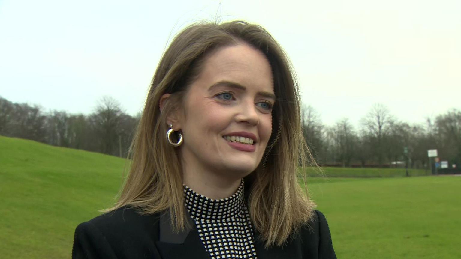 Trása Canavan. She has brown shoulder-length hair and blue eyes, wearing a black and white top and black blazer.