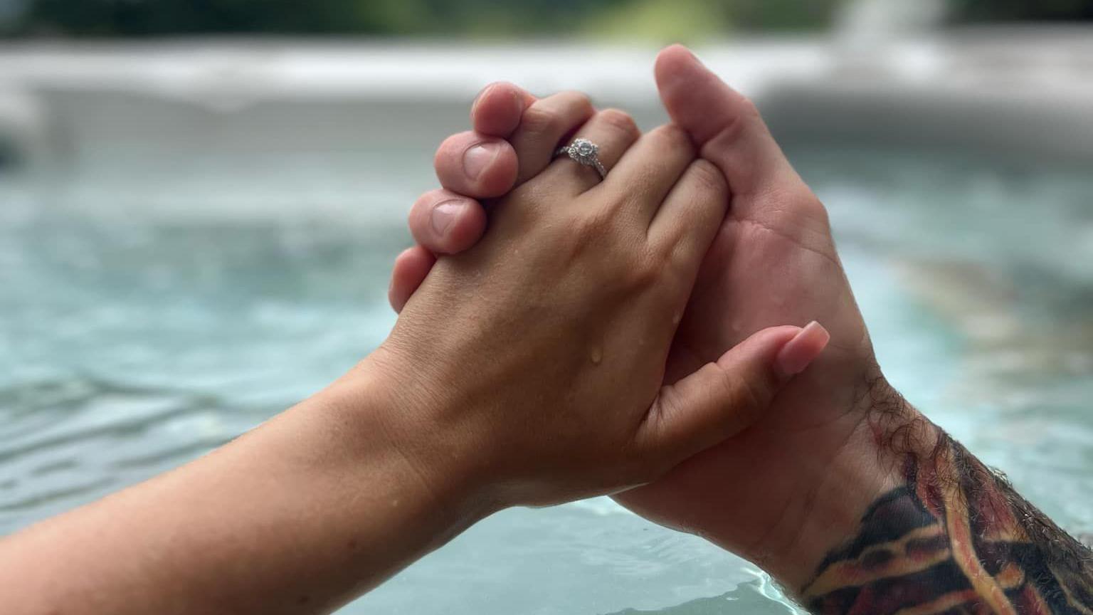 A sleeve tattooed arm, belonging to a man who has just proposed, holds the hand of his fiancée, who is wearing a diamond engagement ring on her finger as they sit in a hot tub