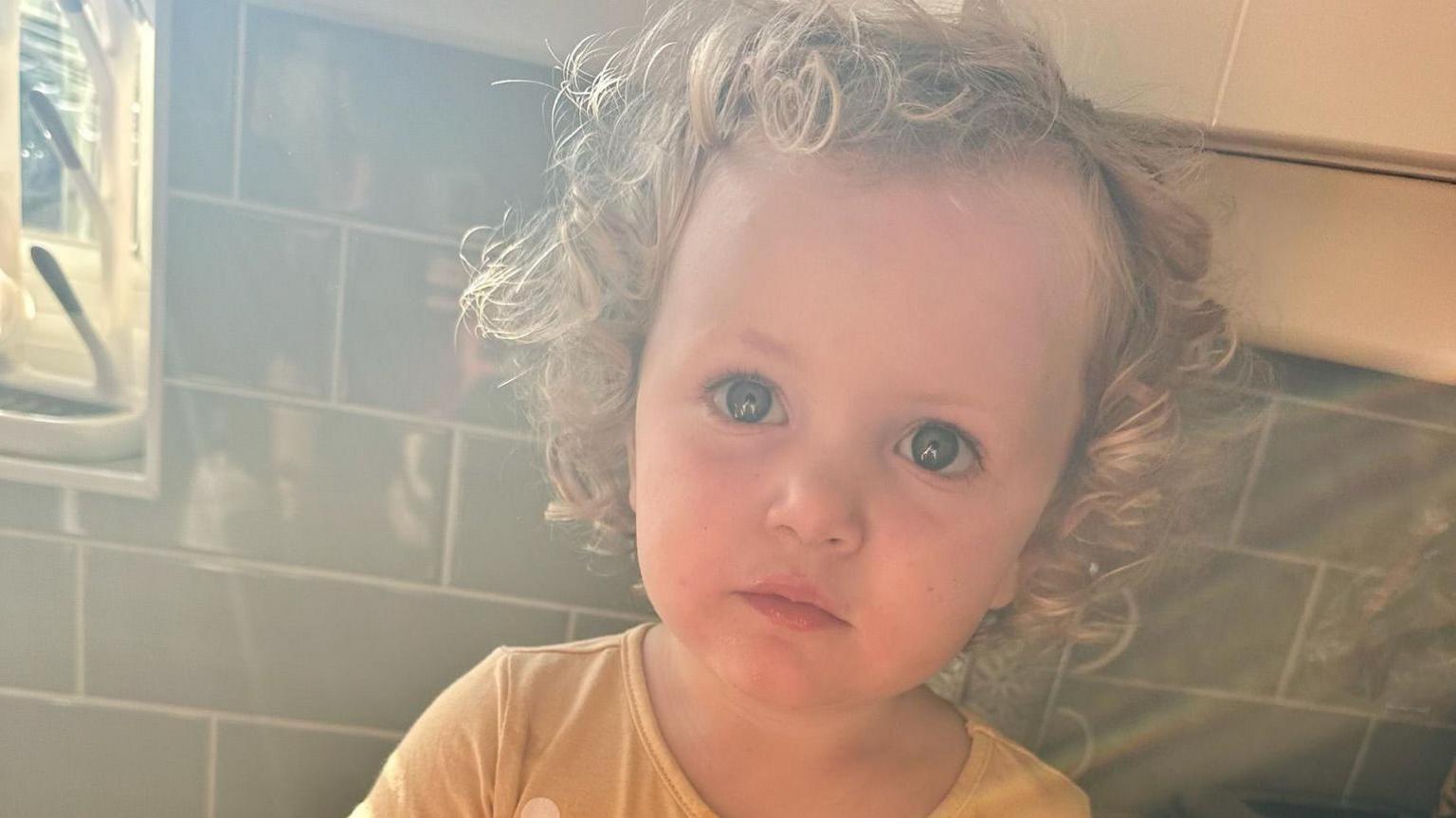 Amber looks at the camera with wide eyes and a serious expression. She has very curly blonde hair and wears a bright yellow top. She appears to be sitting on a kitchen counter, with green tiles behind her.