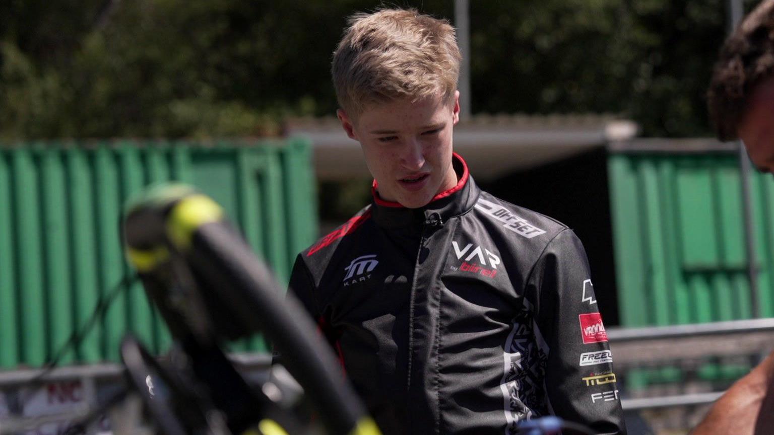 Noah in black racing overalls, watching a car being worked on. He has short light brown hair.