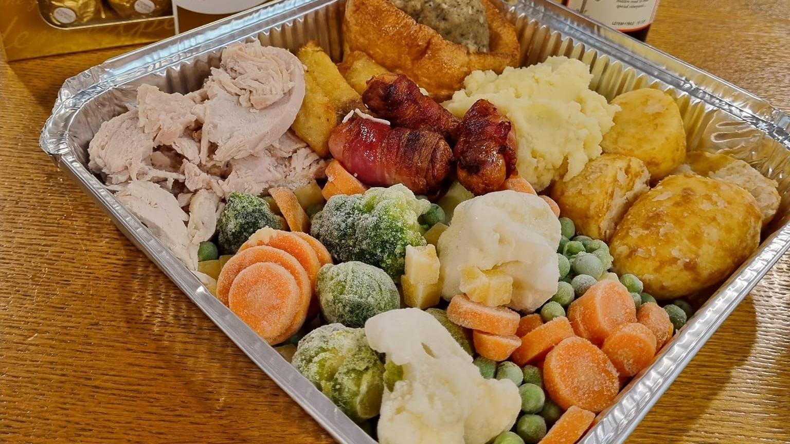 A full roast dinner inside a takeaway metal tray. There is turkey, carrots, broccoli, cauliflower, pigs in blankets, roast potatoes, Yorkshire pudding, stuffing and roast potatoes