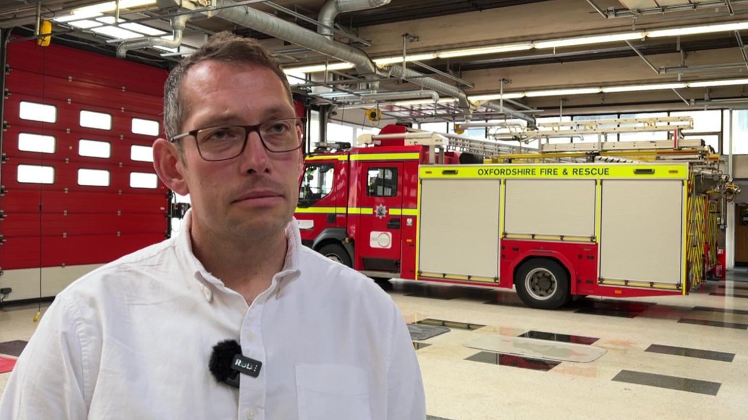 Steve wright has a white shirt on, glasses, and short brown hair. Behind him is a fire station with a fire truck parked in it.