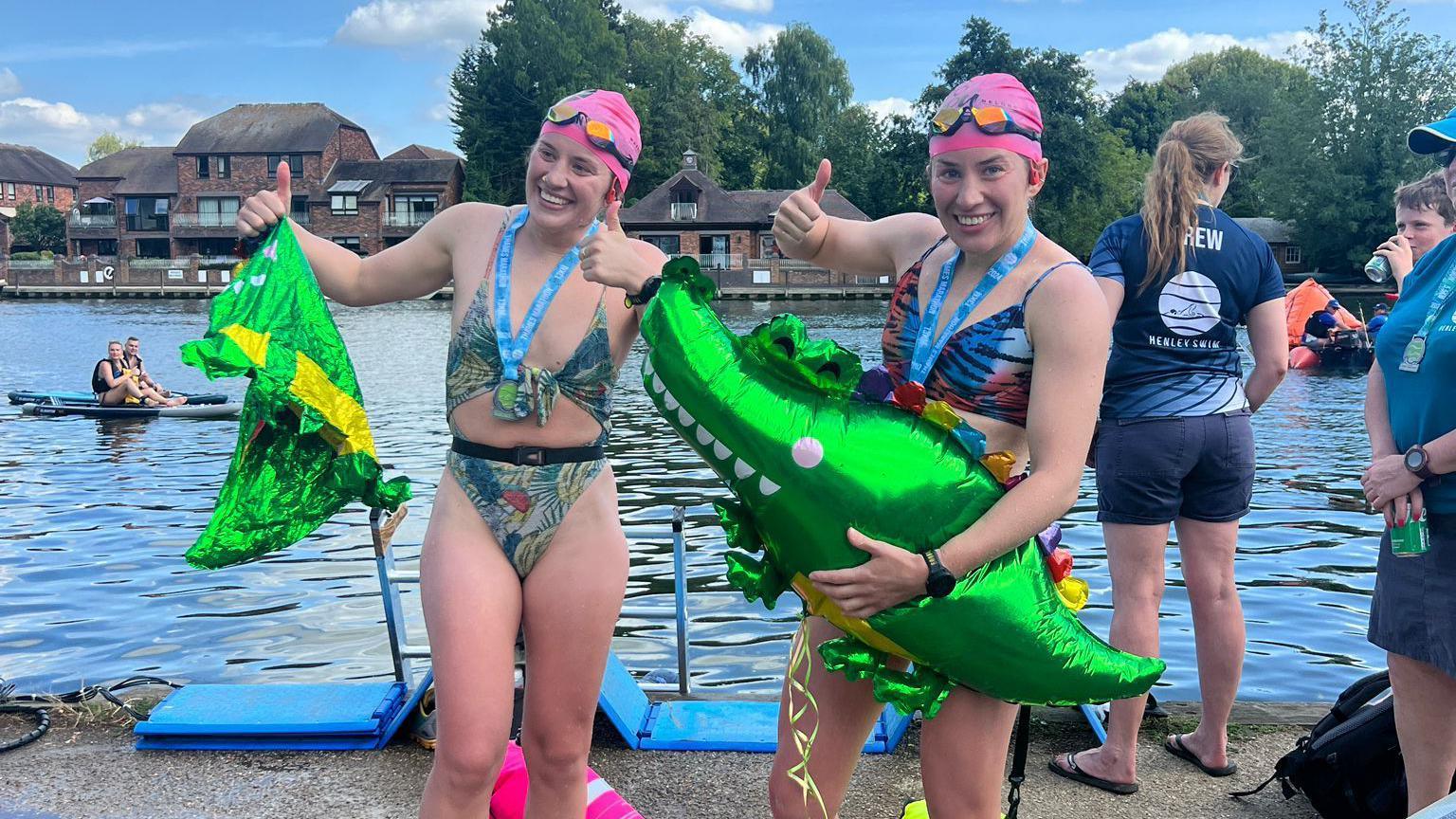 Georgia and Melissa Laurie hold green foil crocodile balloons, wearing swimming costumes and pink swim hats, smiling and have their thumbs up having just completed the swim