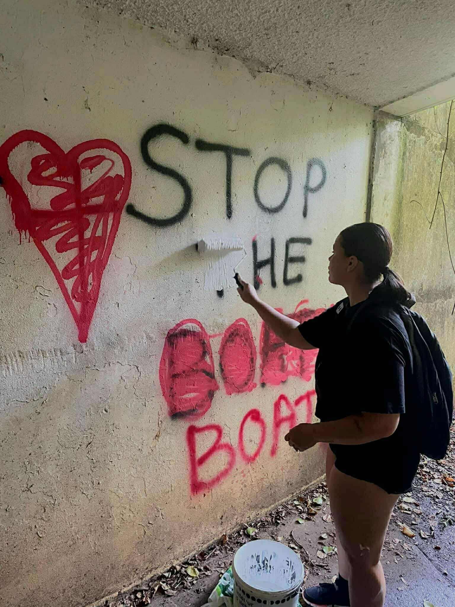 Photograph shows a young woman, wearing a black t-shirt and black shorts, holding a paint brush roller to a subway wall. On the wall, spray painted lettering reads: "Stop he Boats."