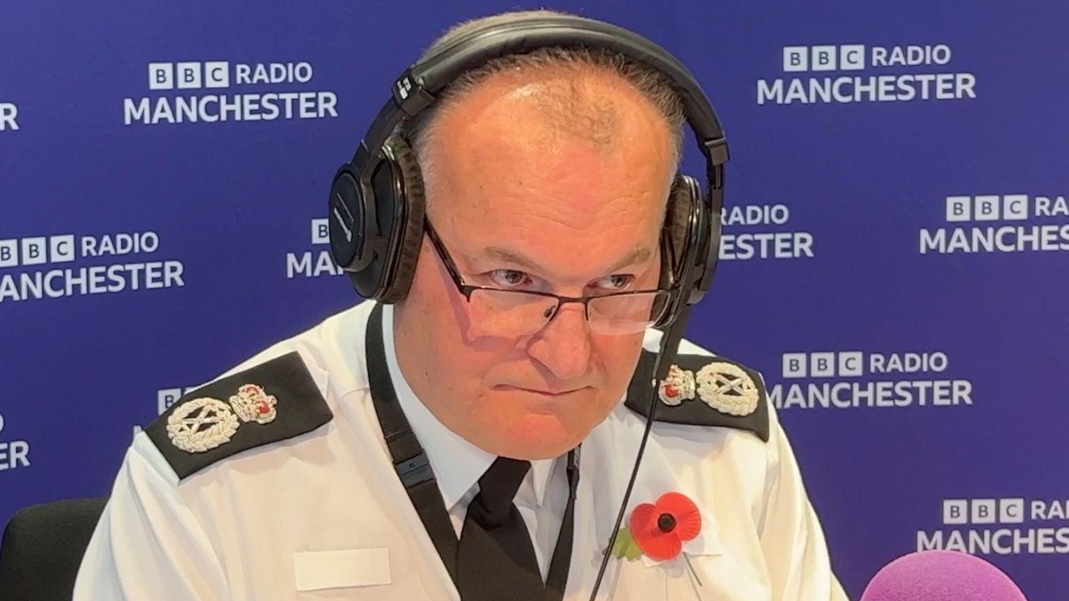 Chief constable Stephen Watson peers over his glasses while wearing black headphones as he takes part in a BBC Radio Manchester phone-in. Behind him can be seen a purple backdrop bearing the BBC Radio Manchester branding. 