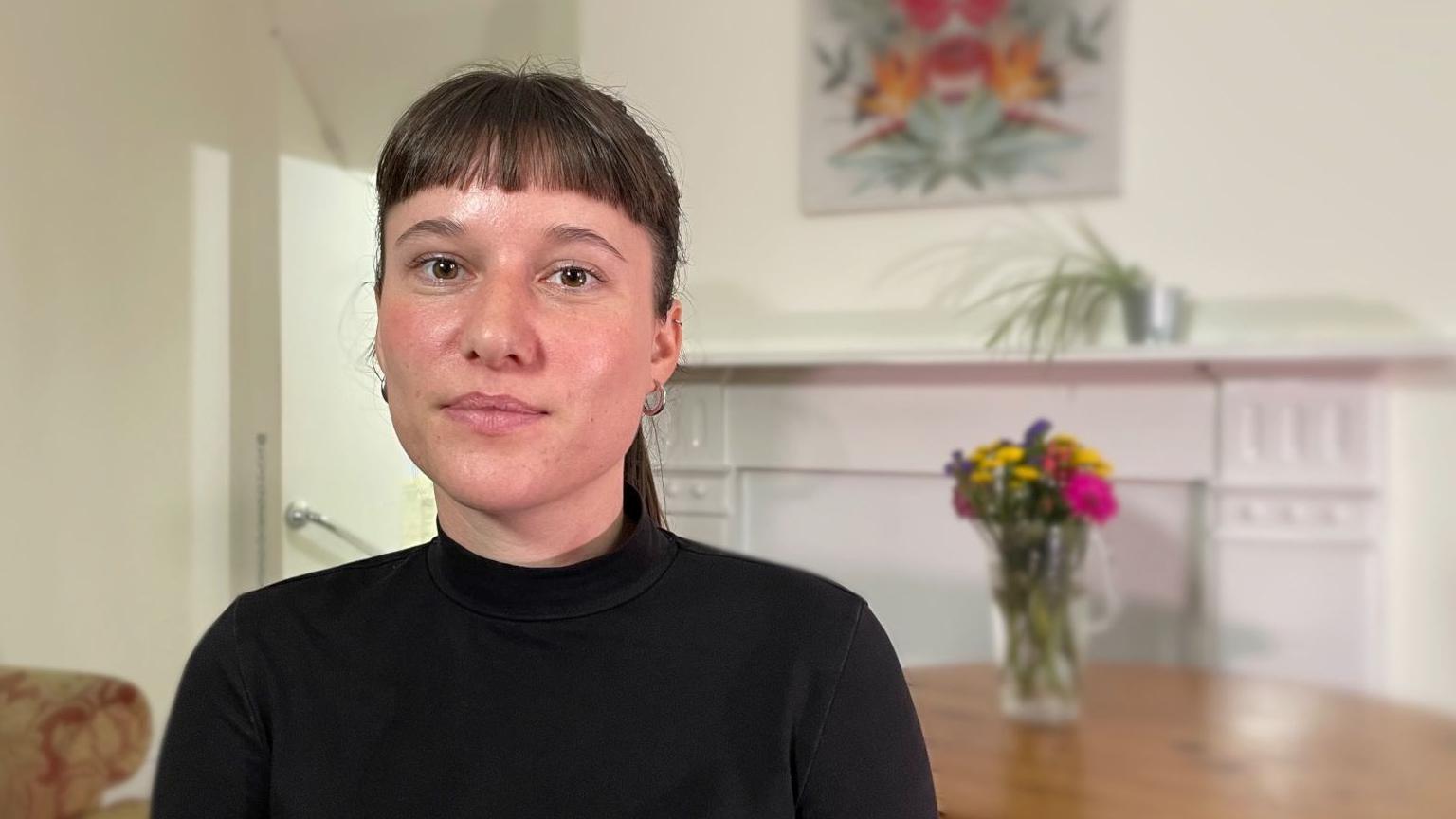 Alice Gilderdale, wearing a black top, small hoop earrings and brown hair in a ponytail, looking directly at the camera. She is seated in front of a small table, which has a jug of flowers on it. In the background is a white fire place surround, with a spider plant in a small pot on the mantelpiece. Above the fire place is an abstract picture. 