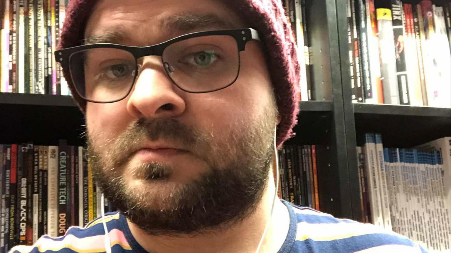 Ben Enser wearing  a red woolly hat and a pink and blue striped T-shirt. There are book cases filled with books behind him. He is wearing glasses and has a brown beard.