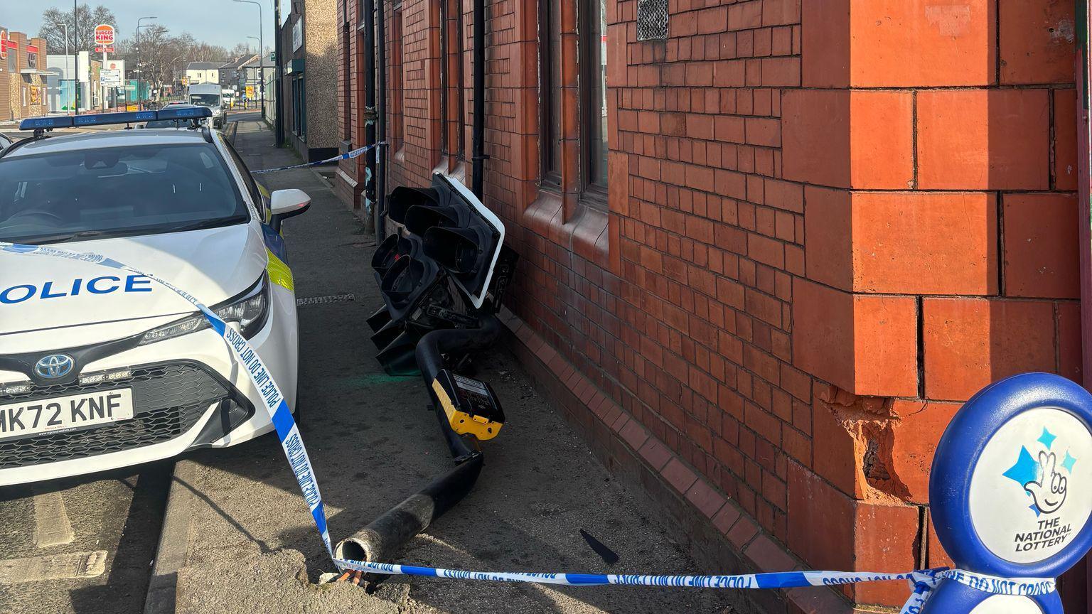 A damaged traffic light lies on the ground next to a police car, police tape hangs over it