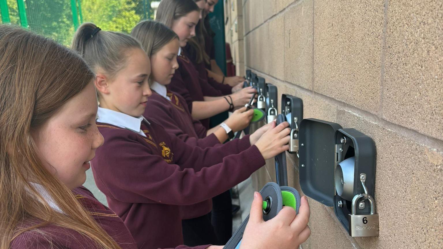 A group of children in red school jumpers locking their phones in boxes
