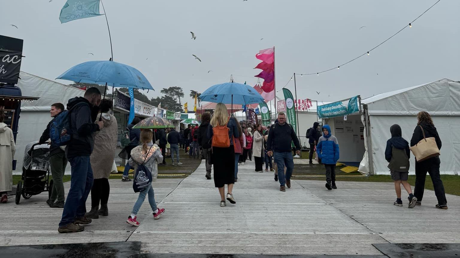 People walking away from the camera carrying blue umbrellas with stalls lining either side of a path. Grey skies can be seen above the stalls. 