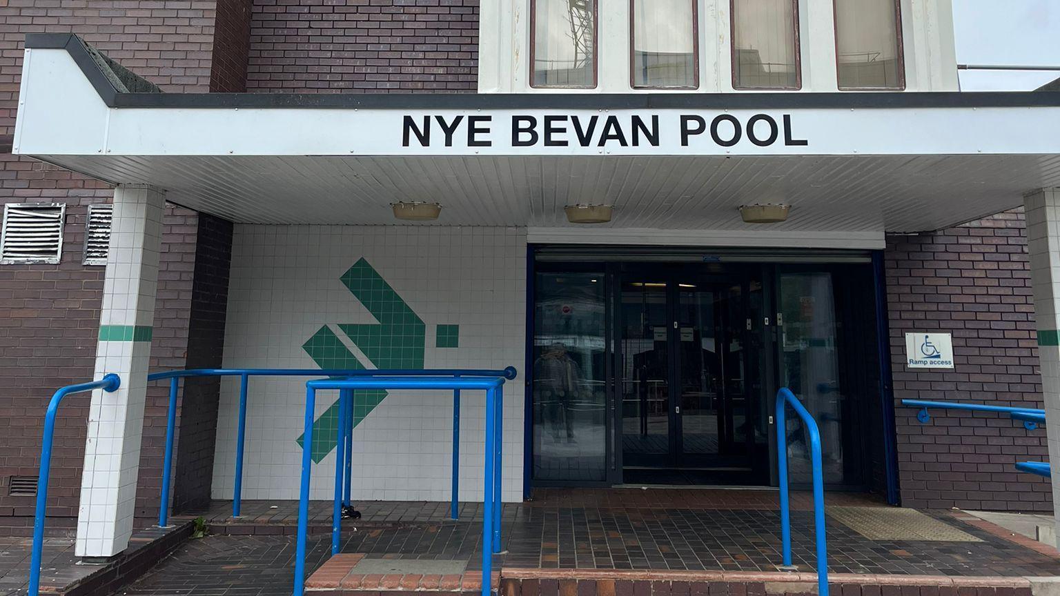 Nye Bevan swimming bath building. It is a dark brown brick building. The front is made up of green and white tiles and automatic glass doors. There are blue railings and also a ramp alongside steps leading up to the entrance.