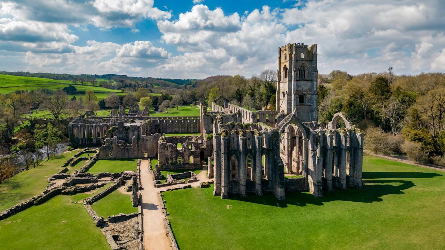 An aerial view of Fountains Abbey in Ripon, with the ruins lit by bright sunlight on a sunny day. 