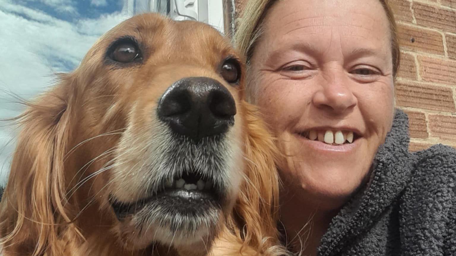 Golden retriever and Irish retriever cross dog with a woman with blonde / brown hair smiling wearing a grey fleece. There is a window and a brick wall behind them.