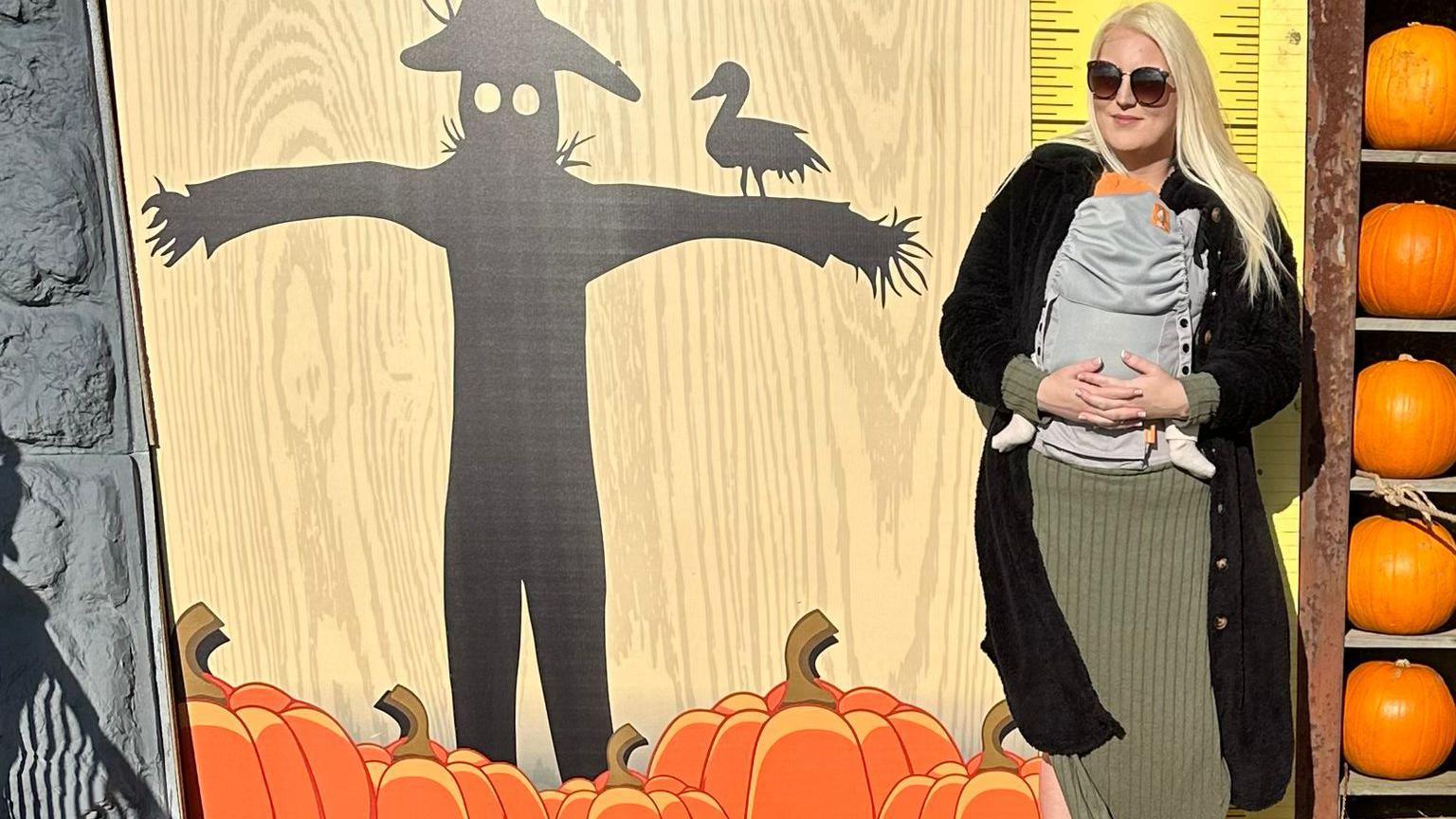 Jodie Fisher standing next to a pumpkin display. She has a baby in her arms and there are pumpkins next to her. 