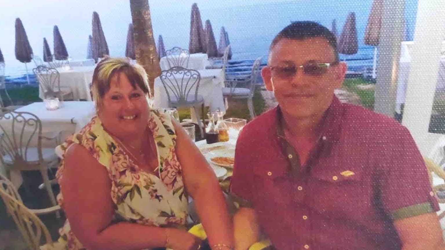 Suzanne and John sit at a table posing for a photo. She is smiling and wearing a flowery blouse - he wears a purple shirt. Appear to be having dinner. 