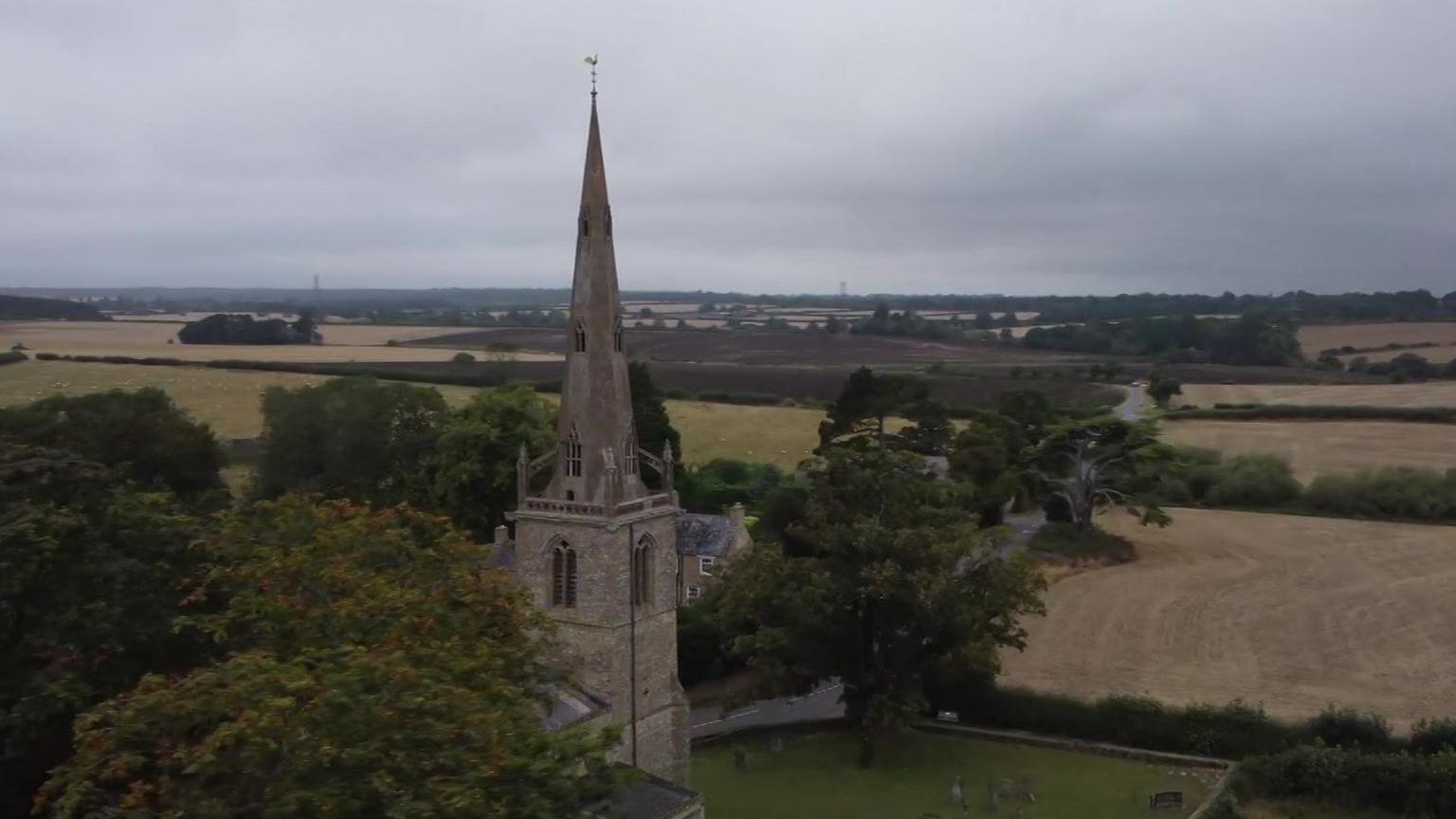 A church with a steeple surrounded by trees and open arable fields.