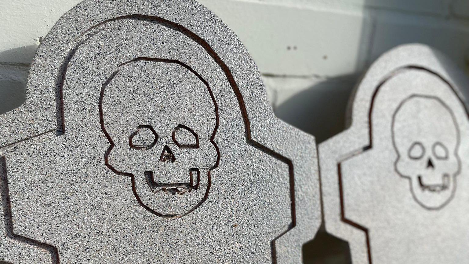 Two grey wooden signs in the shape of headstones. They are leaned against a white wall. They have skulls engraved in them.