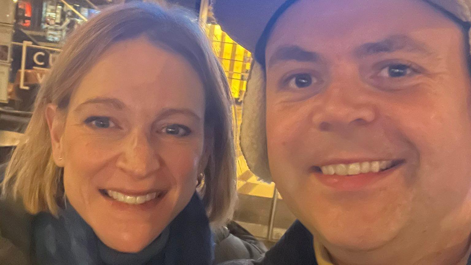 Deb and Geraint smiling at the camera. She has a short blonde bob and he is wearing a cap with ears.