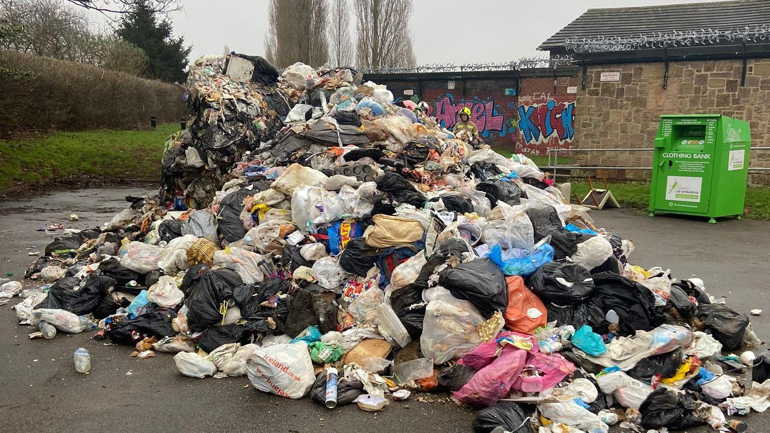 Pile of waste - which had caught fire in a bin lorry - in a car park. A green clothing bank is in the back of the picture towards the right, placed between the pile of rubbish and a stone building.