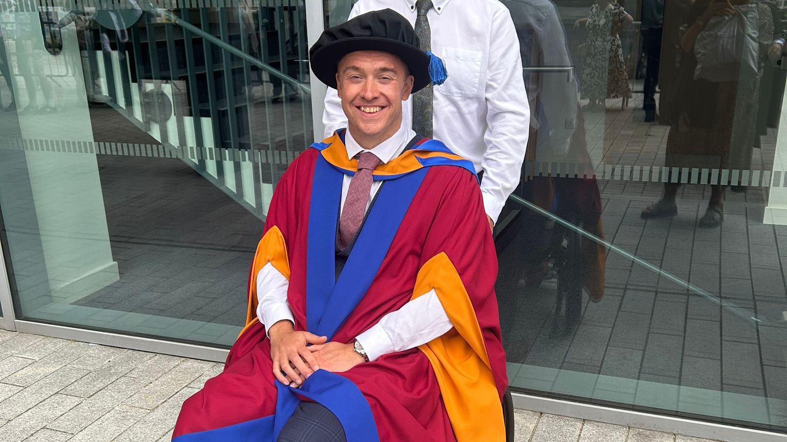 A man in a graduation gown sits in a wheelchair with his hands folded in his lap. He is looking at the camera and smiling.