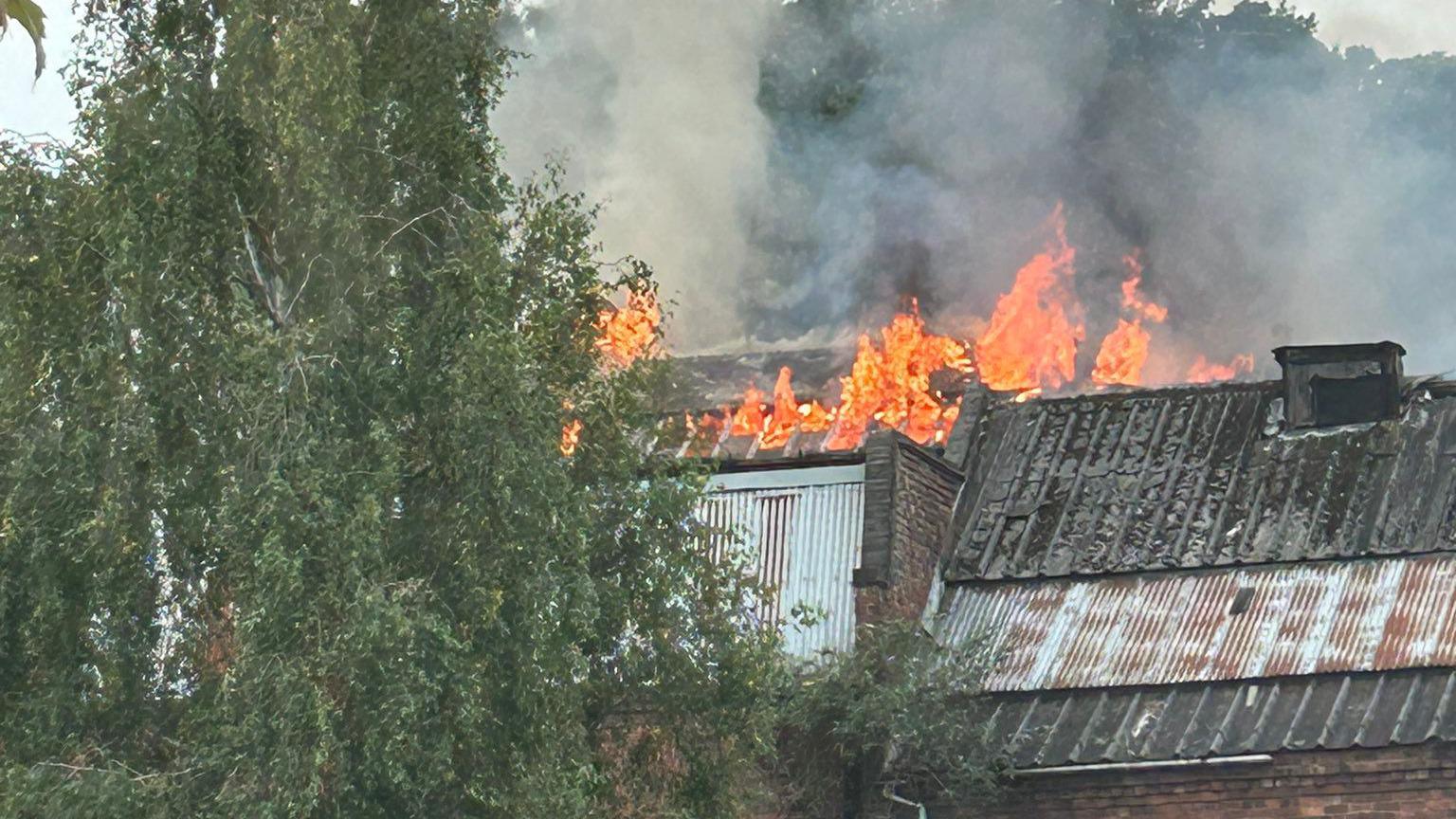 Flames shooting from the roof of a three-story building, with plumes of smoke rising. The building is sited behind a car park.