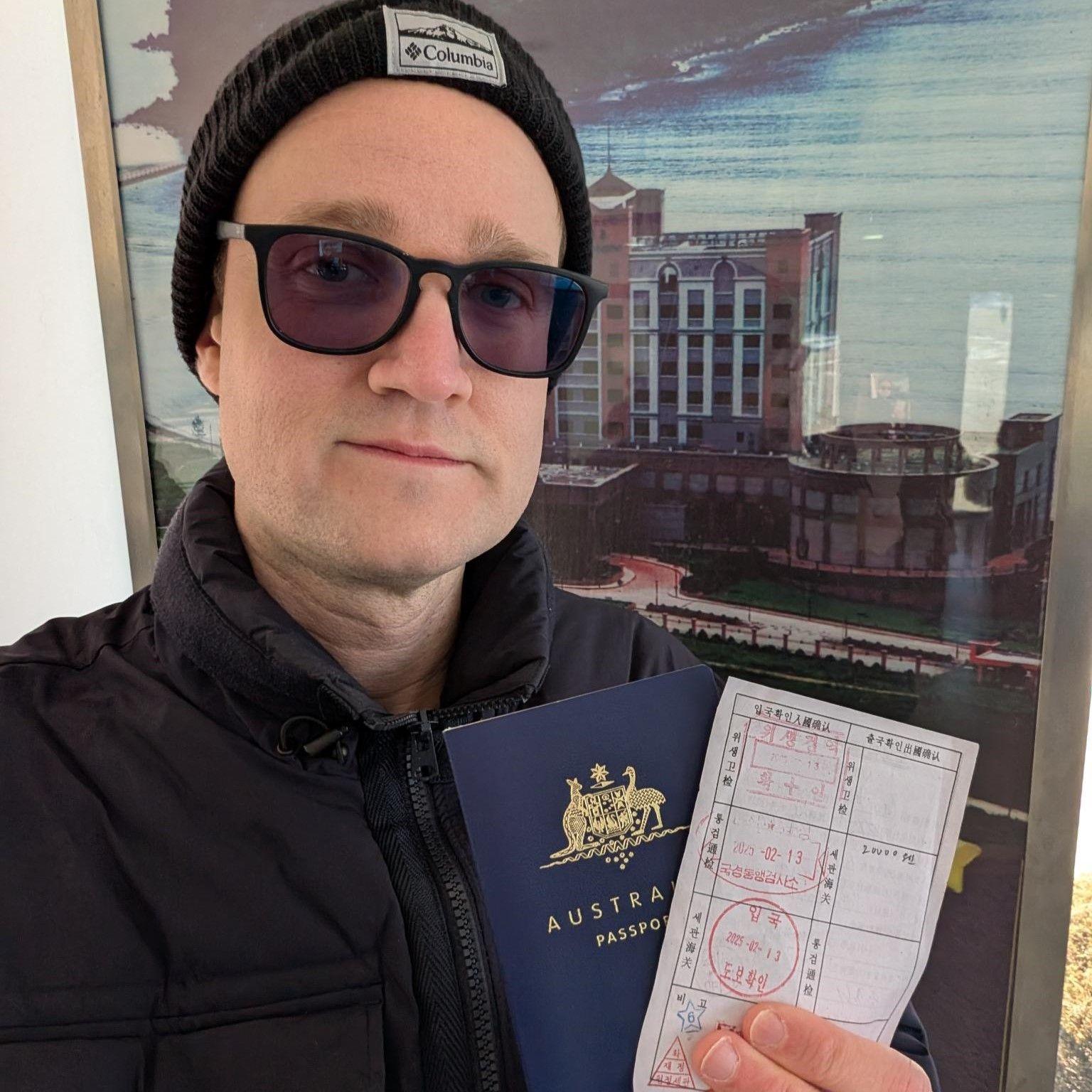 A man in a hat and sunglasses holds a stamped piece of paper and a navy Australian passport