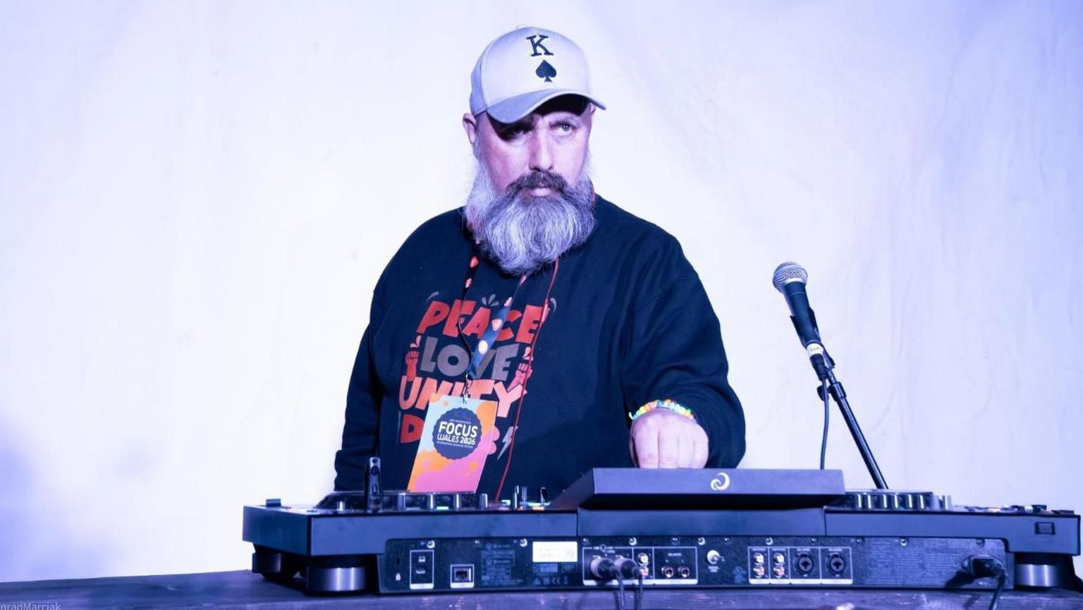 A person wearing a baseball cap and a dark sweatshirt stands behind professional DJ equipment, with one hand on the controls. A microphone on a stand sits to the right, and the scene is lit by bright white and blue-tinted stage lighting against a simple backdrop.
