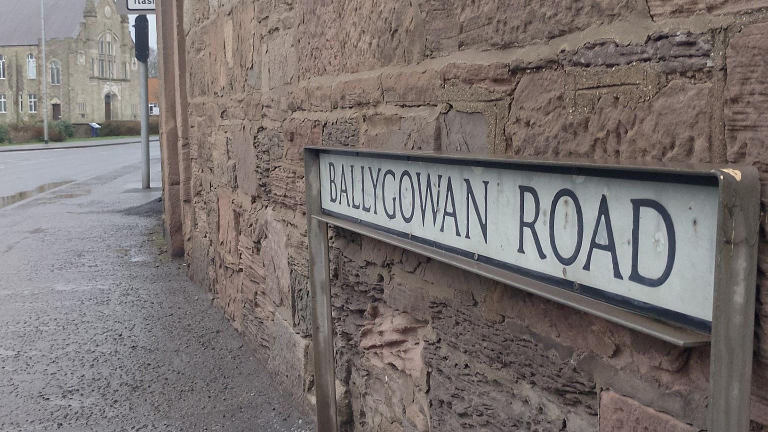 A street corner with the sign "Ballygowan Road" against a red stone wall. The street can be seen behind, and there is a church in the distance. 