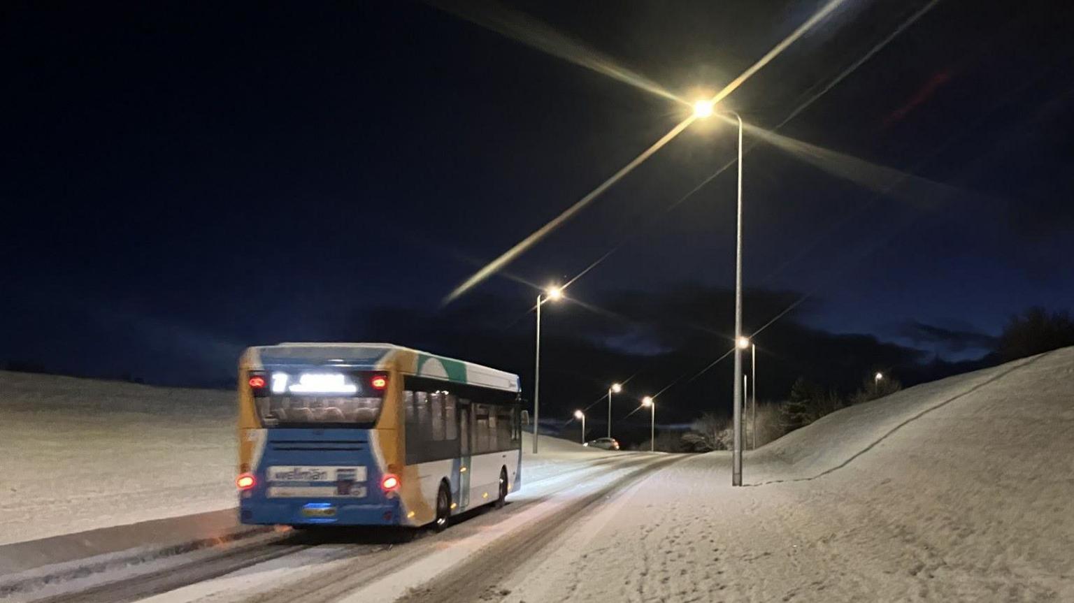 A bus travels along a snow-covered road in Inverness on Wednesday morning. It is still dark and streetlights are on.