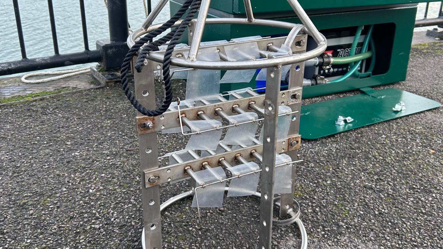 A piece of metal equipment. It is cylindrical in shape. There are thin filters attached to the metal. It is on grey concrete. There is a green box behind it. There is a black fence and blue water behind.