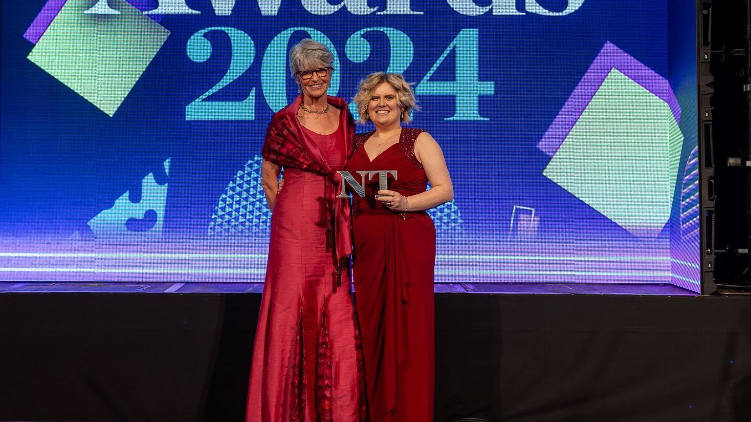 Two women, both wearing red dresses, stand in front of a blue screen. A woman with blonde hair holds a trophy in the shape of two letters, N and T. They are both smiling