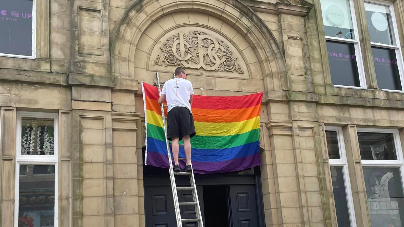 A man is standing at the top of a ladder and is putting up a rainbow flag above the doors of a stone building.