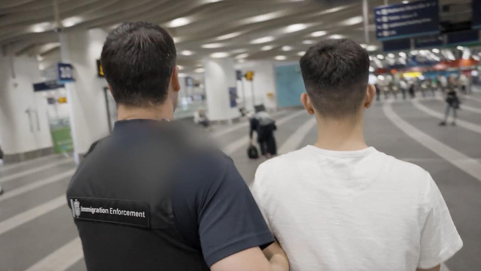 An officer wearing an Immigration Enforcement best his arm hooked under the arm of a man, taking him away. The man looks young but we can only see the backs of their heads. They are in a train station.