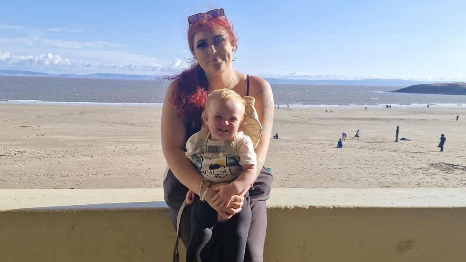 Rebecca Eccleston and her son Theo pictured sitting on concrete wall by beach promenade. The beach and sea are visible in the background. She is wearing black trousers and a black strappy top and has red hair and sunglasses on her head. Theo is smiling and sitting on her lap. He has blonde hair.