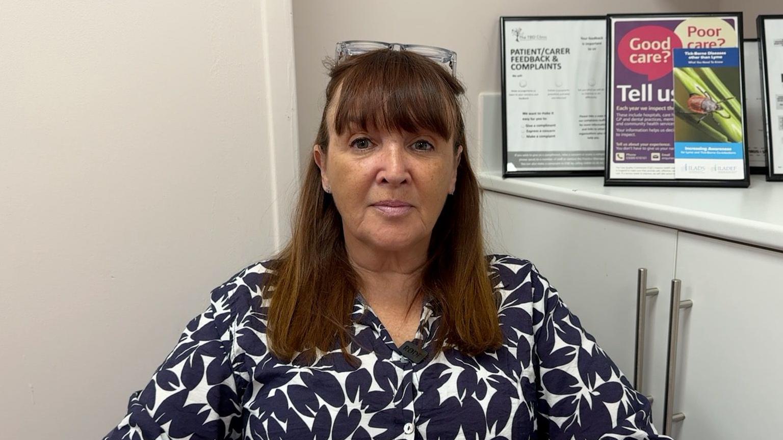 Georgia Tuckey is sat looking at the camera. She is wearing a blue and white shirt, glasses rested on her head and has brown hair. There are diagrams of ticks in the background.