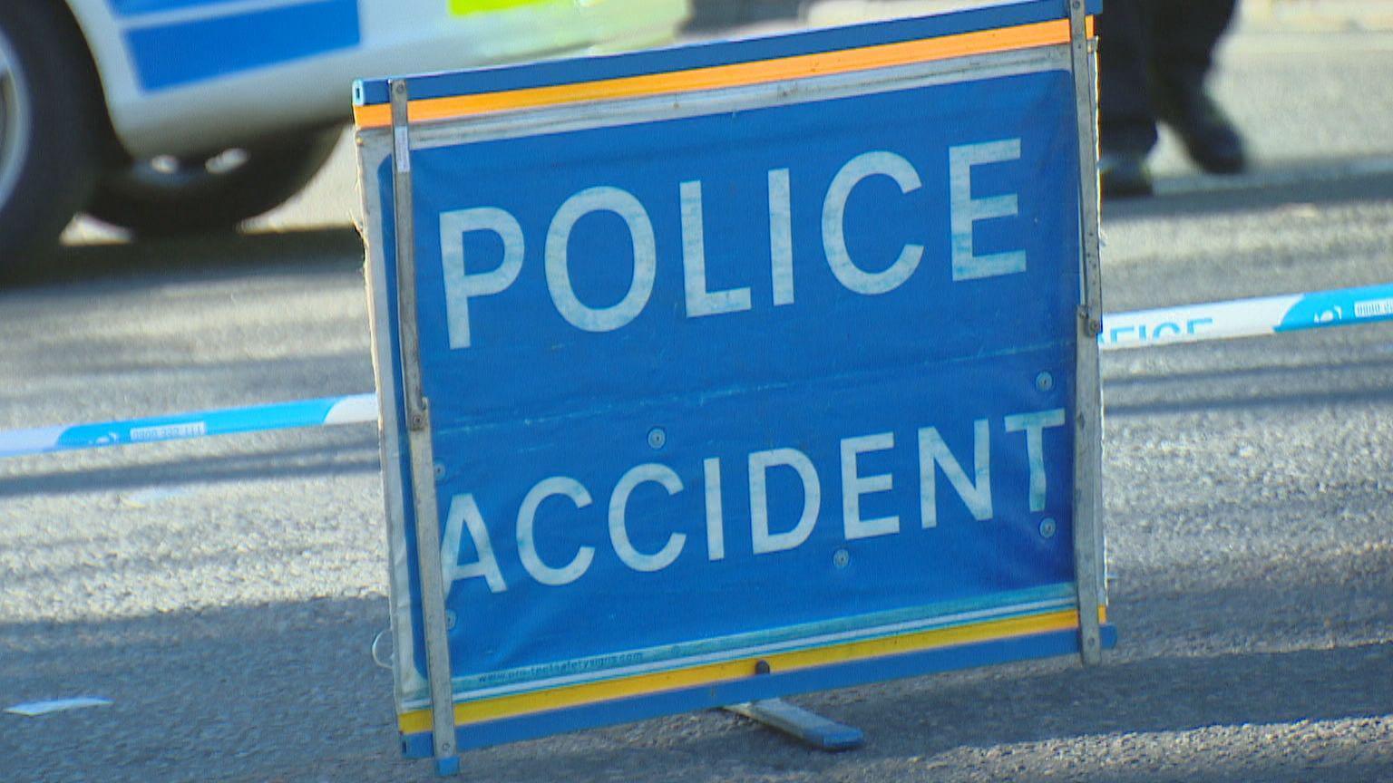 Blue sign with 'police accident' in bold white letters, on a road, with blue and white police tape, and a police vehicle in the background.