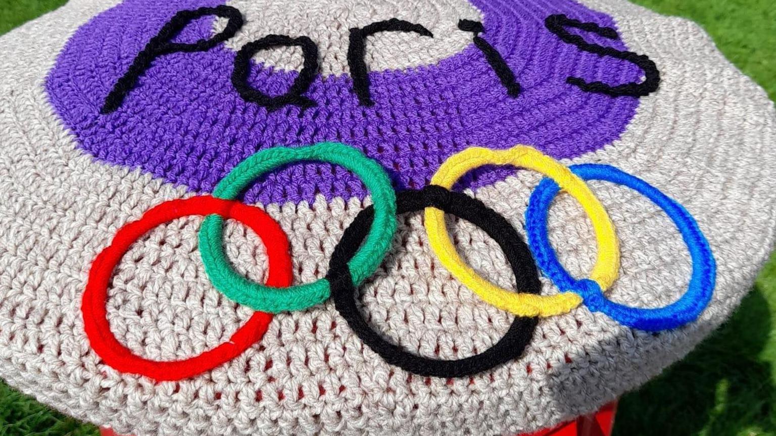 A postbox topper with the word Paris in black against a purple circle and the five Olympic rings below. 