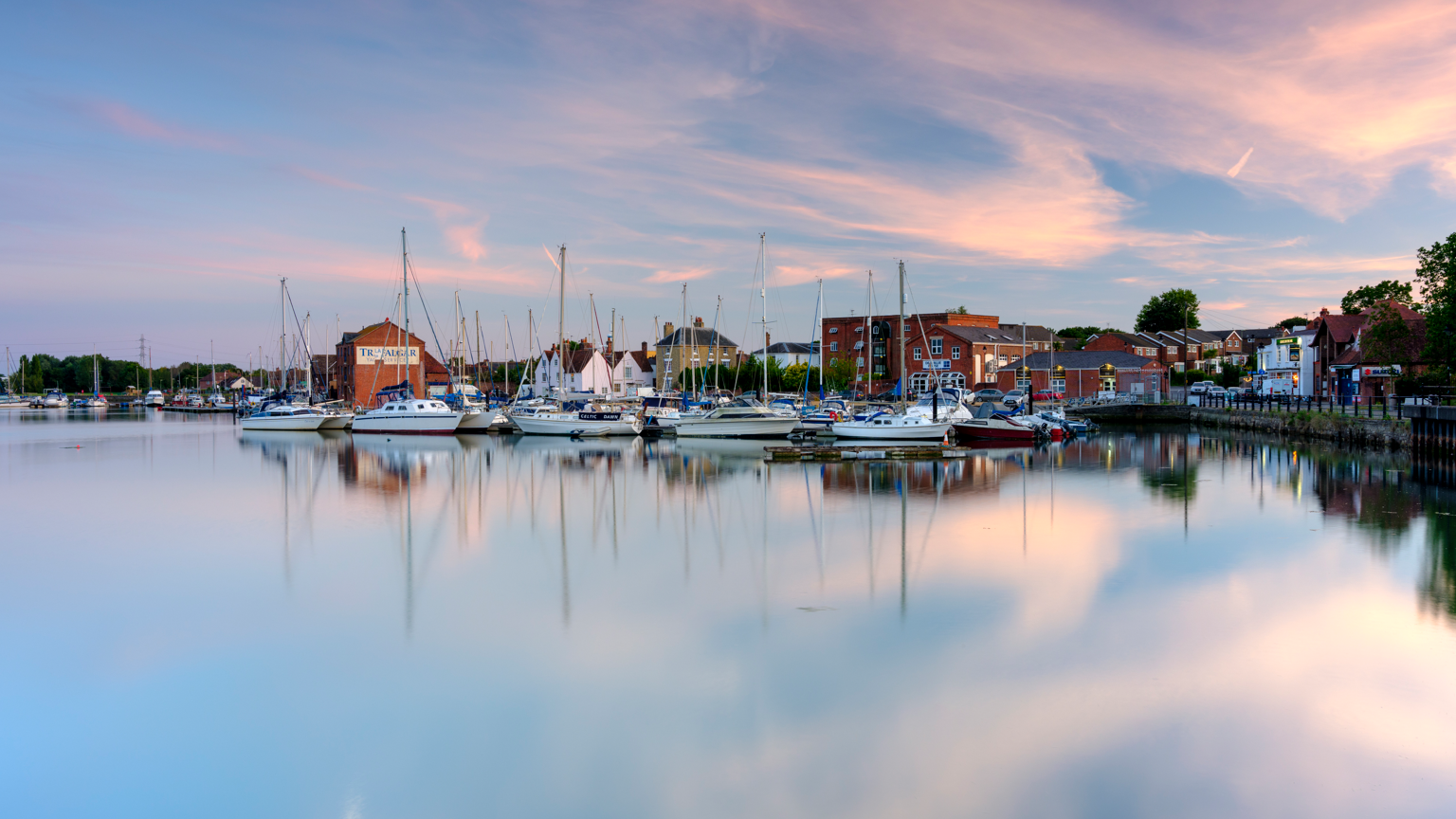 A harbour with small boats in. The sky is pink and blue. There are brick buildings at the edge of the harbour.