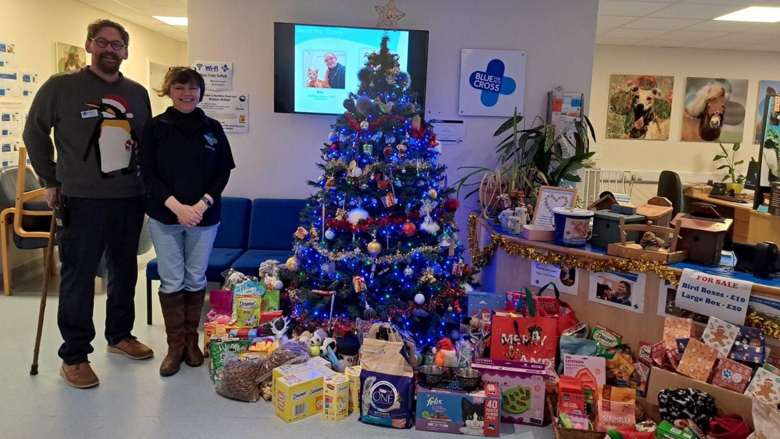 Charles White and a Blue Cross staff member smile at the camera. To the right of them is a Christmas tree surrounded by presents that have been donated for the animals at the centre.