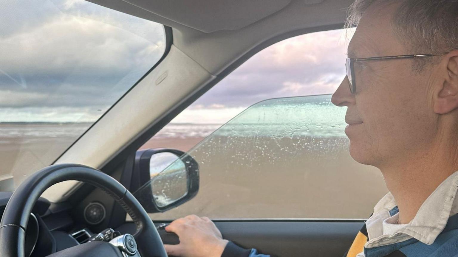 Close-up of profile of Tim Taylor driving his car with the clouds in the background and rain water on the side window.