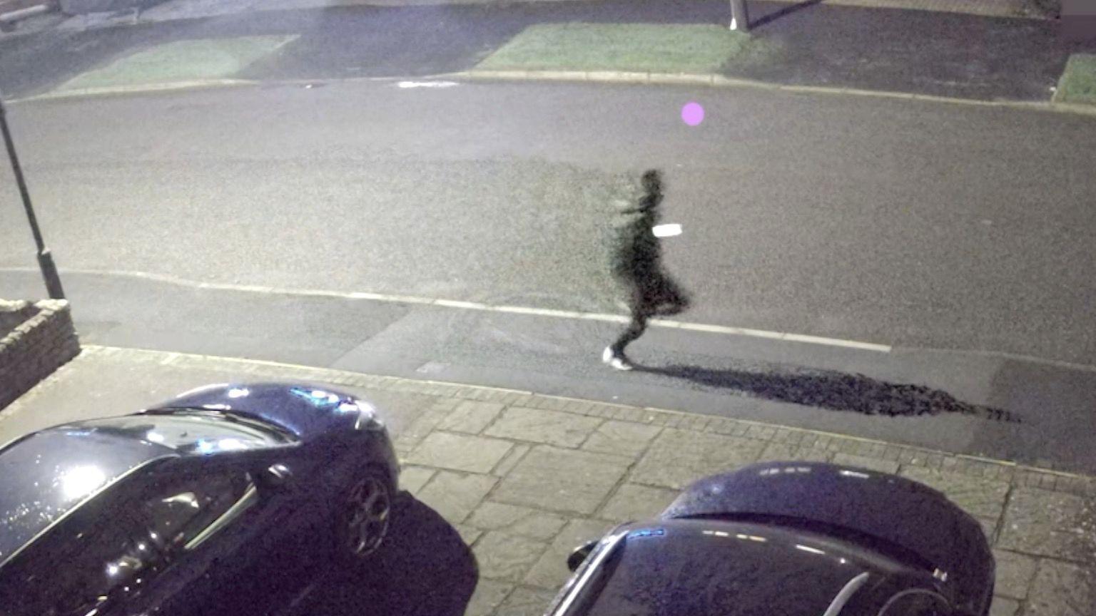 A CCTV image of a dark-clothed person running along a street. The light of a mobile phone can be seen in their hand.
