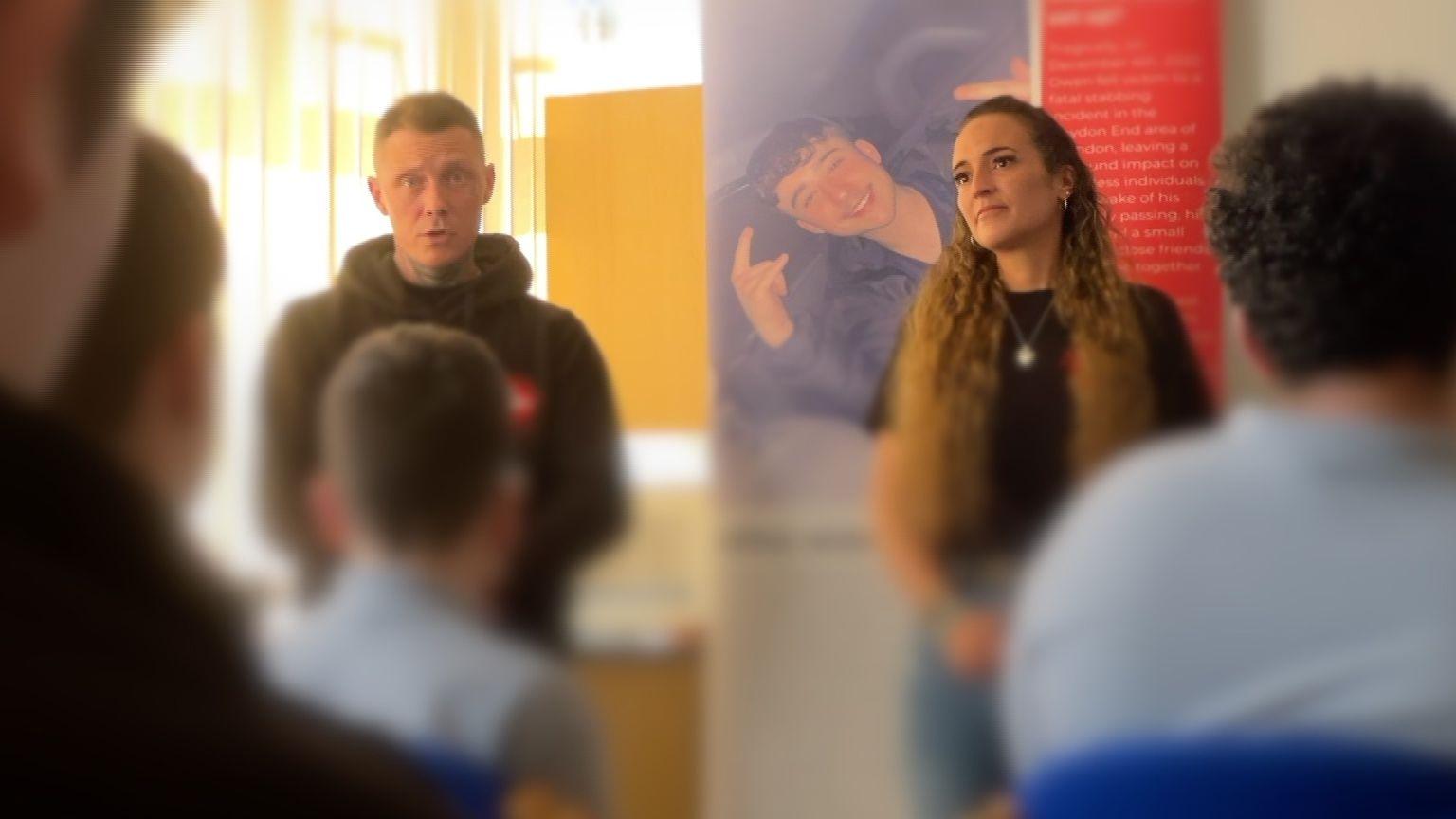 The blurred backs of students facing Danny Hegarty and Jemma Page standing at the front of the class with an Owen's World banner behind them showing Owen Dunn's face