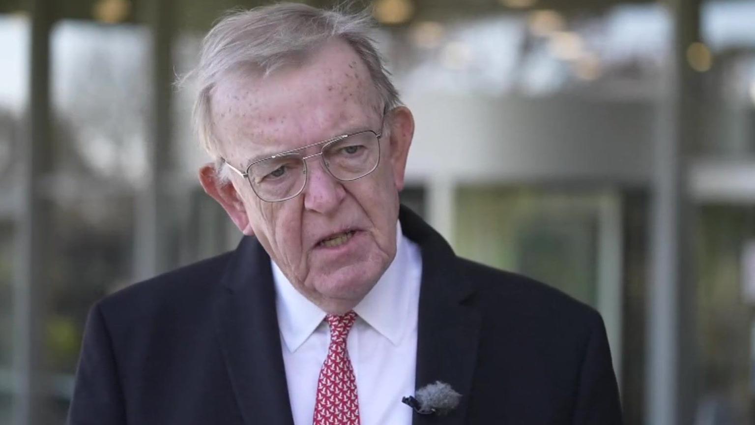 Man with grey hair and glasses wearing dark suit with red tie 